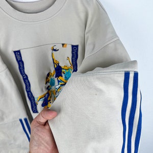 May include: A light gray sweatshirt with blue and white stripes on the sleeves. The sweatshirt has an embroidered image of athletes playing basketball with the text "Olympische Spiele M&uuml;nchen 1972" on the left side and "Olympische" on the right side.