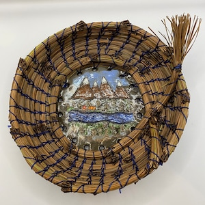 Puede incluir: Una cesta redonda tejida con una baldosa de cerámica azul y marrón que representa una escena de montaña. La cesta está hecha de materiales naturales y tiene un borde azul y blanco.
