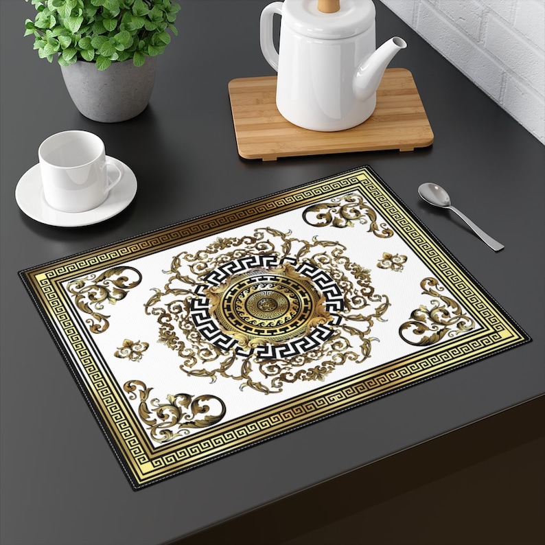 May include: A white placemat with a gold and black geometric design. The design features a large circle with a Greek key pattern and intricate floral details.