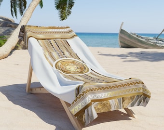 May include: A white beach towel with a gold and brown geometric pattern. The towel is draped over a wooden beach chair on a sandy beach.
