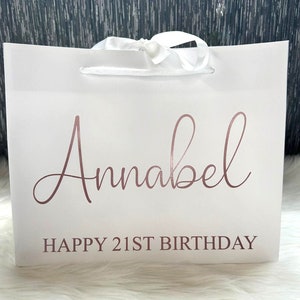 May include: White gift bag with a white satin ribbon and the text "Annabel Happy 21st Birthday" in rose gold lettering.