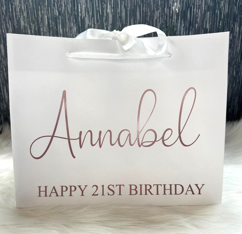May include: White gift bag with a white satin ribbon and the text "Annabel Happy 21st Birthday" in rose gold lettering.