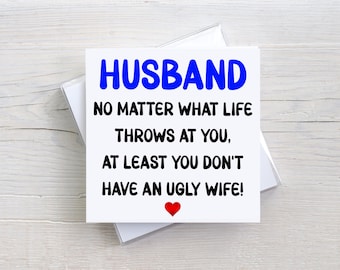 May include: A square white greeting card with blue text that reads HUSBAND and black text that reads NO MATTER WHAT LIFE THROWS AT YOU, AT LEAST YOU DON'T HAVE AN UGLY WIFE! with a small red heart icon centered at the bottom of the card.