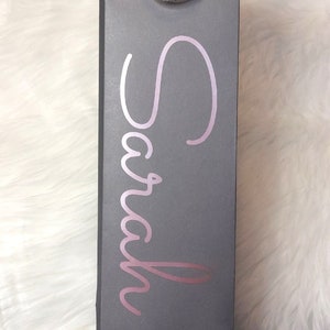 May include: Gray wine bottle gift bag with a silver handle. The name "Sarah" is written in a cursive, rose-gold font on the front of the bag. The bag is rectangular and stands upright.