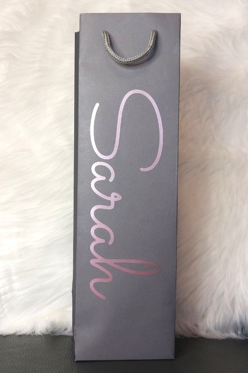 May include: Gray wine bottle gift bag with a silver handle. The name "Sarah" is written in a cursive, rose-gold font on the front of the bag. The bag is rectangular and stands upright.