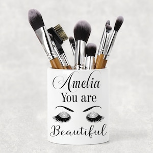 May include: White ceramic makeup brush holder with the text "Amelia You are Beautiful" and a pair of stylised eyes with eyelashes and glitter. The holder is filled with various makeup brushes.