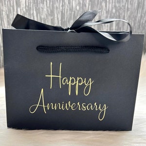 May include: Black gift bag with a black ribbon and gold lettering that says "Happy Anniversary".