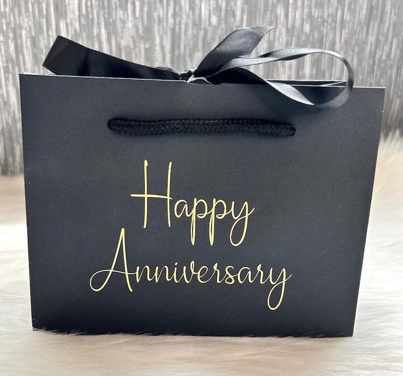 May include: Black gift bag with a black ribbon and gold lettering that says "Happy Anniversary".