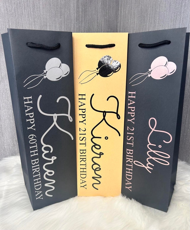 May include: Three gift bags with balloon graphics and personalized text. The bags are black and tan, with the names Karen, Kieron, and Lilly printed in silver and pink. The text includes "Happy 60th Birthday" and "Happy 21st Birthday".