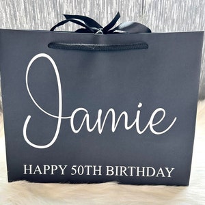 May include: Black gift bag with a white satin ribbon and a white rope handle. The bag is personalized with the name "Jamie" and the text "Happy 50th Birthday".