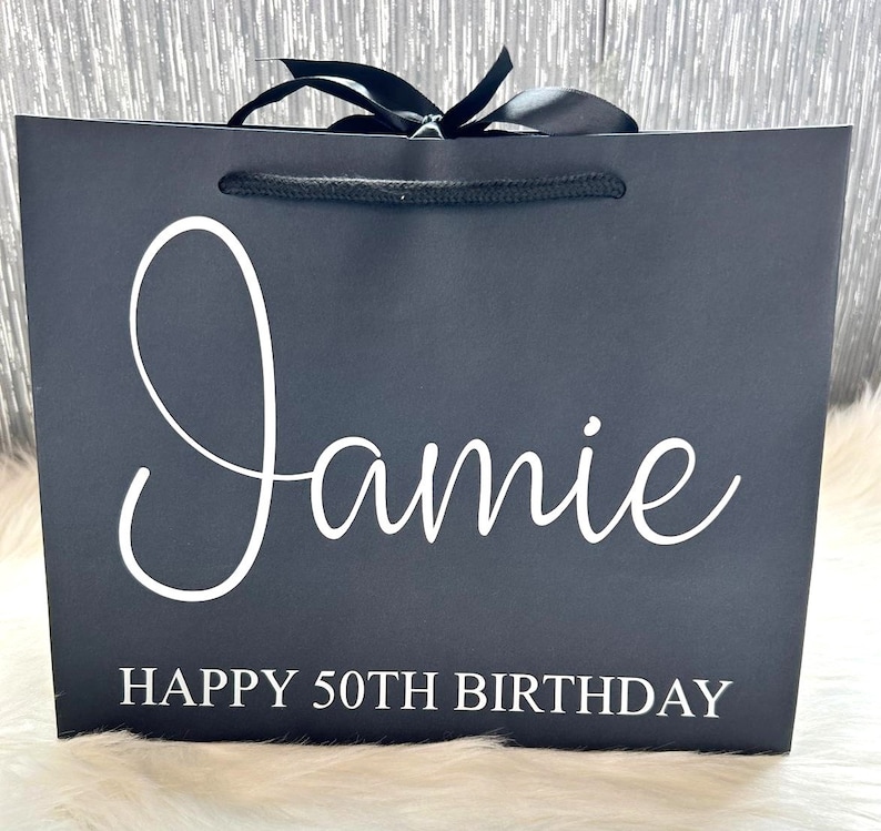 May include: Black gift bag with a white satin ribbon and a white rope handle. The bag is personalized with the name "Jamie" and the text "Happy 50th Birthday".