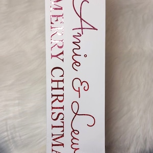 May include: White gift bag with a red glittery "Merry Christmas" and "Annie & Lewis" text. The bag has a white cord handle and is designed for a bottle. The background is a white fluffy surface.