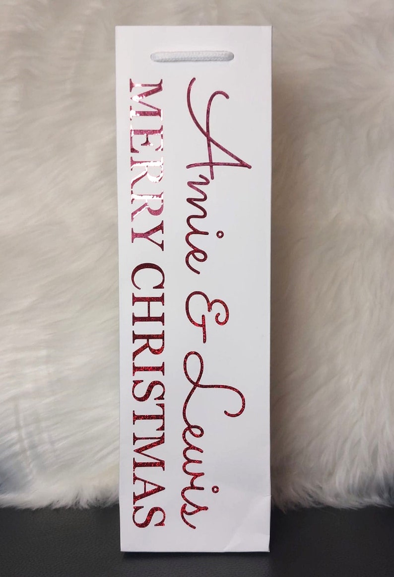 May include: White gift bag with a red glittery "Merry Christmas" and "Annie & Lewis" text. The bag has a white cord handle and is designed for a bottle. The background is a white fluffy surface.