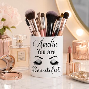 May include: A white cylindrical makeup brush holder featuring the black printed text Amelia You are Beautiful and a graphic of closed eyes with glittery eyelashes. The holder is filled with various makeup brushes with metallic handles.