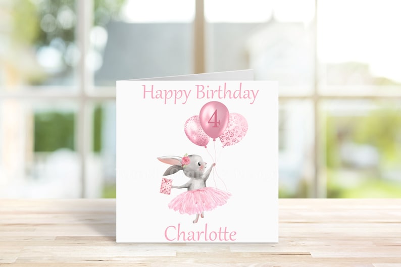 May include: A pink birthday card featuring a grey bunny wearing a pink tutu and holding pink balloons. One balloon has a pink "4" on it, while the others have a pink floral pattern. The card reads "Happy Birthday Charlotte".