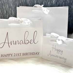 May include: Three white gift bags with satin ribbon bows. The largest bag is blank. The middle bag has the text "Annabel Happy 21st Birthday" in rose gold lettering. The smallest bag has the text "Happy Anniversary" in silver lettering.
