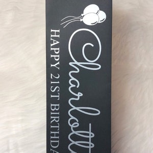 May include: Black wine bottle gift bag with a black cord handle. The bag features the name "Charlotte" in a cursive font, along with the text "HAPPY 21ST BIRTHDAY" and a graphic of three balloons in white.