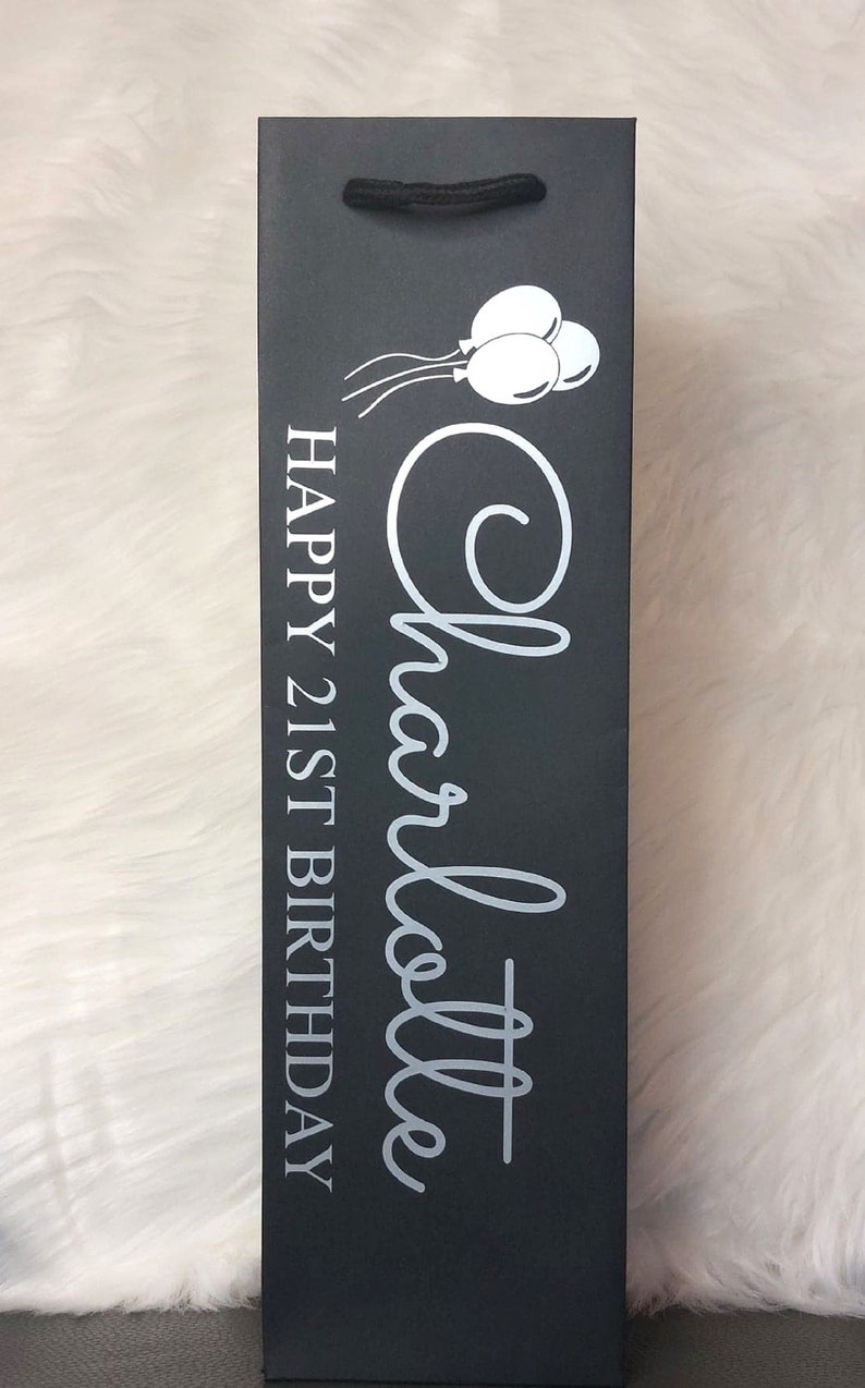 May include: Black wine bottle gift bag with a black cord handle. The bag features the name "Charlotte" in a cursive font, along with the text "HAPPY 21ST BIRTHDAY" and a graphic of three balloons in white.