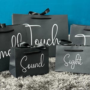 May include: Five black gift bags with white lettering. The bags are arranged in a pyramid shape. The bags are labelled "Smell", "Touch", "Sound", "Sight", and "Taste".