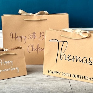 K&ouml;nnte beinhalten: Drei braune Papiert&uuml;ten mit wei&szlig;er und schwarzer Beschriftung. Die gr&ouml;&szlig;te T&uuml;te sagt "Happy 30th Birthday Chelsea". Die mittlere T&uuml;te sagt "Thomas HAPPY 26TH BIRTHDAY". Die kleinste T&uuml;te sagt "Happy Anniversary".
