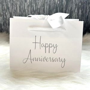 May include: White gift bag with a white satin ribbon and the text "Happy Anniversary" printed in gray.