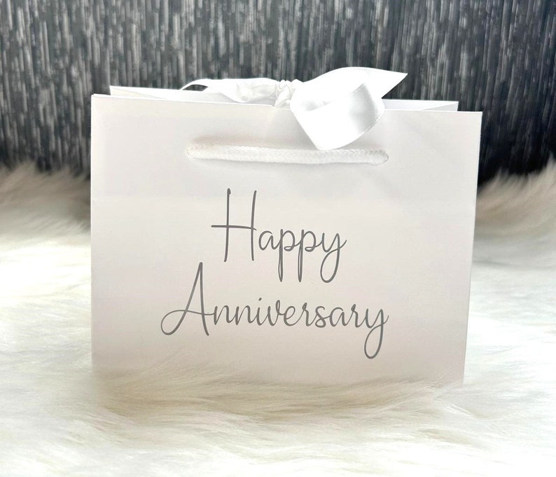 May include: White gift bag with a white satin ribbon and the text "Happy Anniversary" printed in gray.