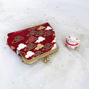 Peut inclure: Une bourse à monnaie en tissu rouge et or avec un motif de nuages et un fermoir en métal doré. La bourse est posée sur un fond blanc avec des paillettes dorées.
