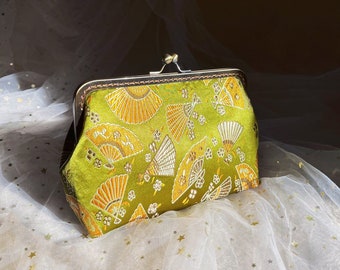 Puede incluir: Un bolso de mano de tela verde con cierre de plata y un diseño floral con abanicos. El bolso está sobre un fondo de tul blanco.