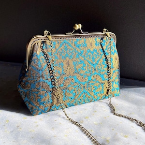 Puede incluir: Un bolso de mano con estampado turquesa y dorado con una correa de cadena. El bolso presenta un cierre decorativo y un diseño floral detallado. La cadena es dorada y el bolso está sobre una superficie blanca.
