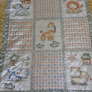 May include: A white baby quilt with a patchwork design featuring various animals, including a zebra, giraffe, lion, elephant, and hippopotamus. The quilt has a yellow and green border with a checkered pattern.