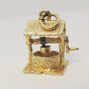May include: A gold charm shaped like a miniature wishing well, featuring a textured stone-like base, a wooden-style roof, a small crank handle on the side, and a tiny bucket suspended by a dark cord from the central beam.