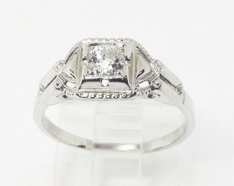 May include: A vintage engagement ring with a single round diamond set in a square, ornate, platinum setting. The band is plain and has a delicate milgrain edge.