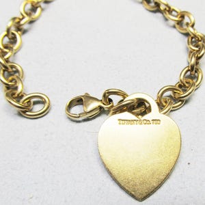 May include: A gold-toned bracelet with a heart-shaped charm. The charm is engraved with "TIFFANY & CO. 750". The bracelet features a chain link design and a clasp closure. The jewelry is likely made of gold.