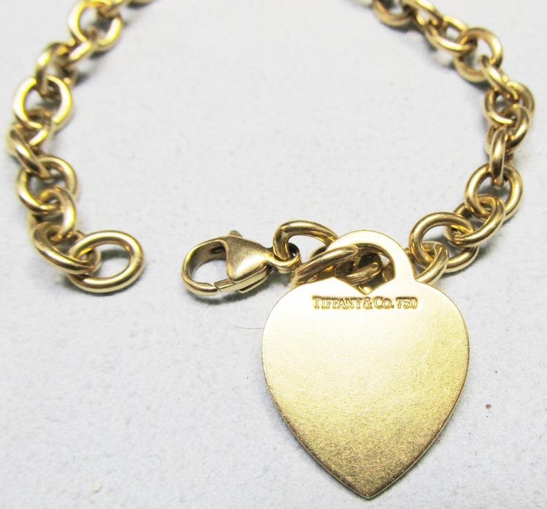 May include: A gold-toned bracelet with a heart-shaped charm. The charm is engraved with "TIFFANY & CO. 750". The bracelet features a chain link design and a clasp closure. The jewelry is likely made of gold.