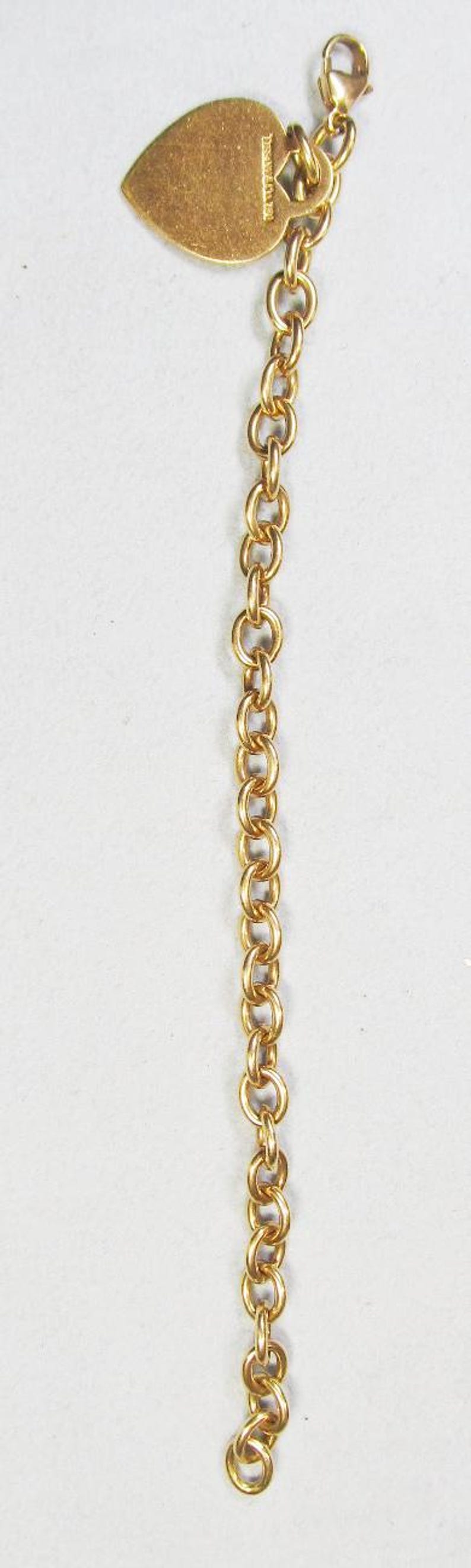 May include: Gold-toned chain with a heart-shaped charm. The charm has a smooth surface and appears to have engraved text. The chain is made of small, interlocking oval links and has a lobster clasp.