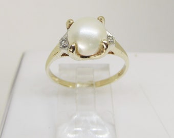 May include: A gold ring featuring a large, oval-shaped pearl as the centerpiece. The pearl is a creamy white color and is held in place by four gold prongs. Small accent diamonds are set on either side of the pearl. The ring is displayed on a clear stand.