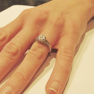 May include: A silver ring with a delicate filigree design and a single round diamond in the center.