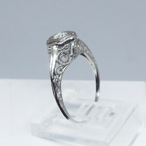 May include: A delicate silver ring with a round, clear gemstone set in a filigree design. The ring is on a white display stand.