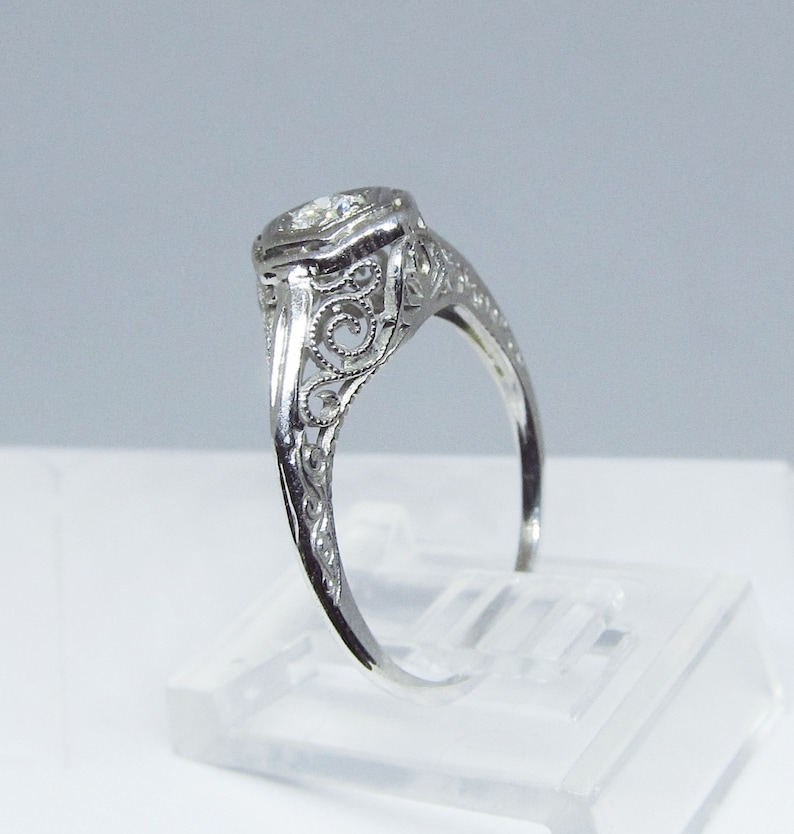 May include: A delicate silver ring with a round, clear gemstone set in a filigree design. The ring is on a white display stand.