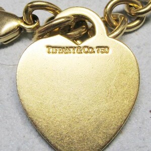 May include: Close-up of a gold-colored heart-shaped charm bracelet. The charm is engraved with "TIFFANY & CO. 750". The bracelet features a lobster clasp and a chain link design. The charm has a brushed metal finish.
