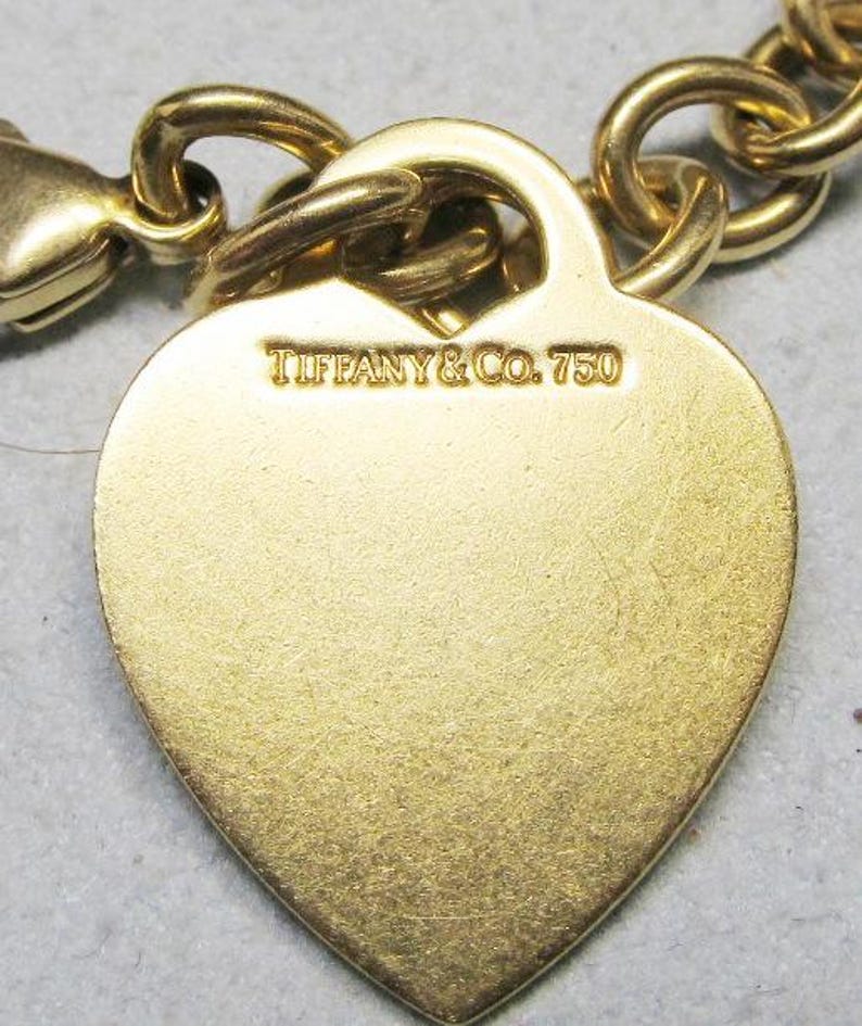 May include: Close-up of a gold-colored heart-shaped charm bracelet. The charm is engraved with "TIFFANY & CO. 750". The bracelet features a lobster clasp and a chain link design. The charm has a brushed metal finish.