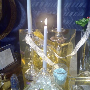 Puede incluir: Tres velas blancas encendidas en candelabros de plata con una cinta dorada y blanca. Las velas están sobre una mesa con un cuenco de vidrio transparente lleno de cristales transparentes y un frasco de vidrio transparente lleno de cristales azules.