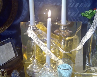 May include: Three white candles are lit on silver candle holders with a gold and white ribbon. The candles are on a table with a clear glass bowl filled with clear crystals and a clear glass jar filled with blue crystals.