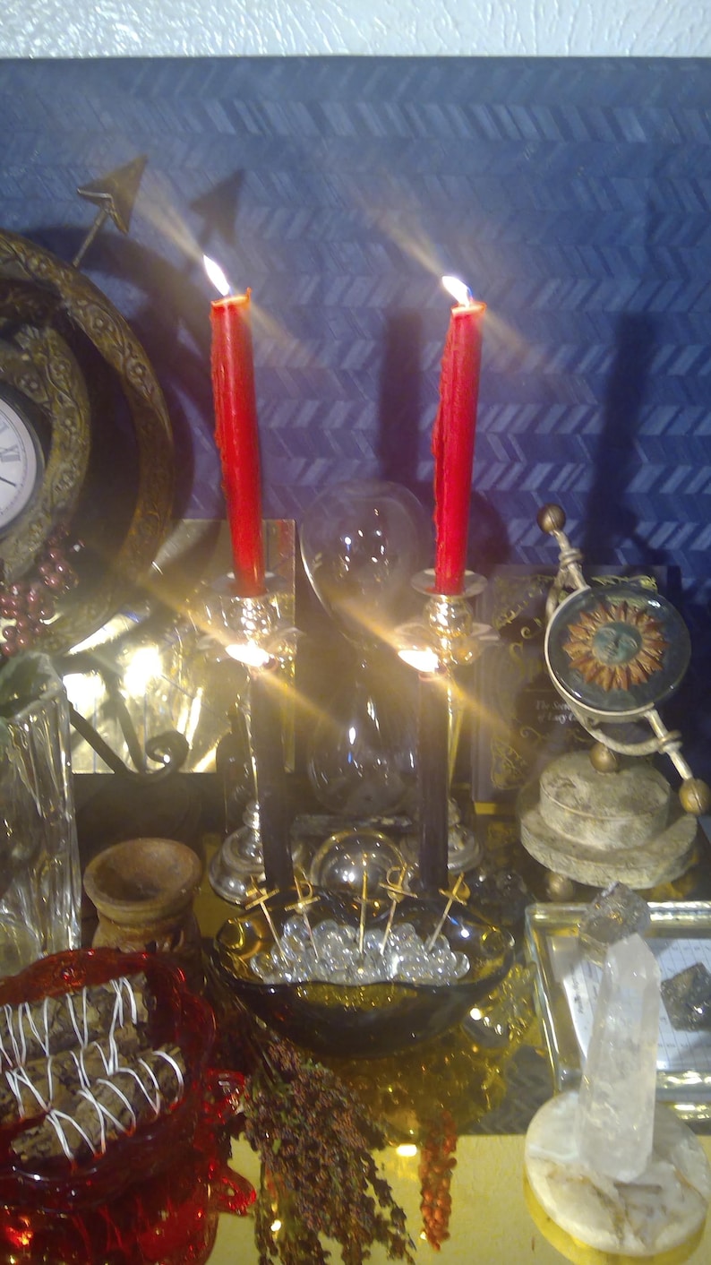 May include: Two red candles are lit in gold candle holders on a table with a variety of decorative items, including a glass orb, a small wooden bowl, and a crystal on a gold stand.