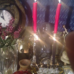 May include: A still life featuring two lit red candles, a glass vase with red berries and greenery, a glass bowl with clear crystals and gold metal accents, and a red glass bowl with white sage smudge sticks.