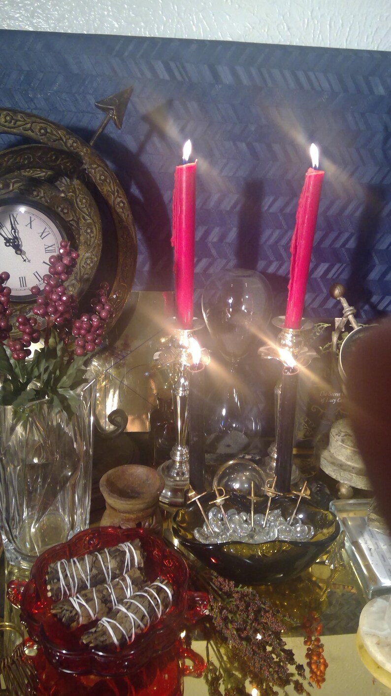 May include: A still life featuring two lit red candles, a glass vase with red berries and greenery, a glass bowl with clear crystals and gold metal accents, and a red glass bowl with white sage smudge sticks.
