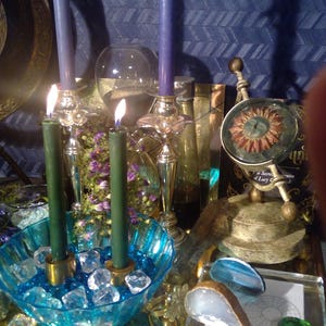 May include: A blue and green candle magic altar with a blue glass bowl filled with clear crystals, a gold metal stand with a round top and three legs, a clear glass bottle, a small gold metal stand with a round top and a glass orb with a red sun design, and a blue and white agate slice.