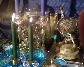 May include: A blue and green candle magic altar with a blue glass bowl filled with clear crystals, a gold metal stand with a round top and three legs, a clear glass bottle, a small gold metal stand with a round top and a glass orb with a red sun design, and a blue and white agate slice.