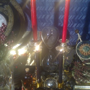 Może przedstawiać: Zbliżenie na świecznik z dwoma zapalonymi czerwonymi świecami. Świecznik jest wykonany z metalu i ma czarne wykończenie. Świece znajdują się w szklanej misce ze złotą obwódką. W tle znajduje się klepsydra i inne elementy dekoracyjne.