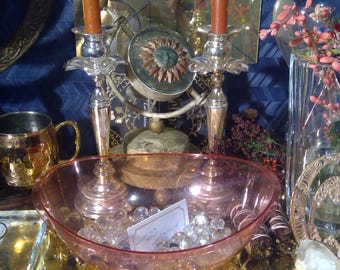 May include: A pink glass bowl filled with clear glass beads and a small white card with the text "Wildwood" sits on a table with two silver candle holders with lit orange candles, a copper mug, and other decorative items.